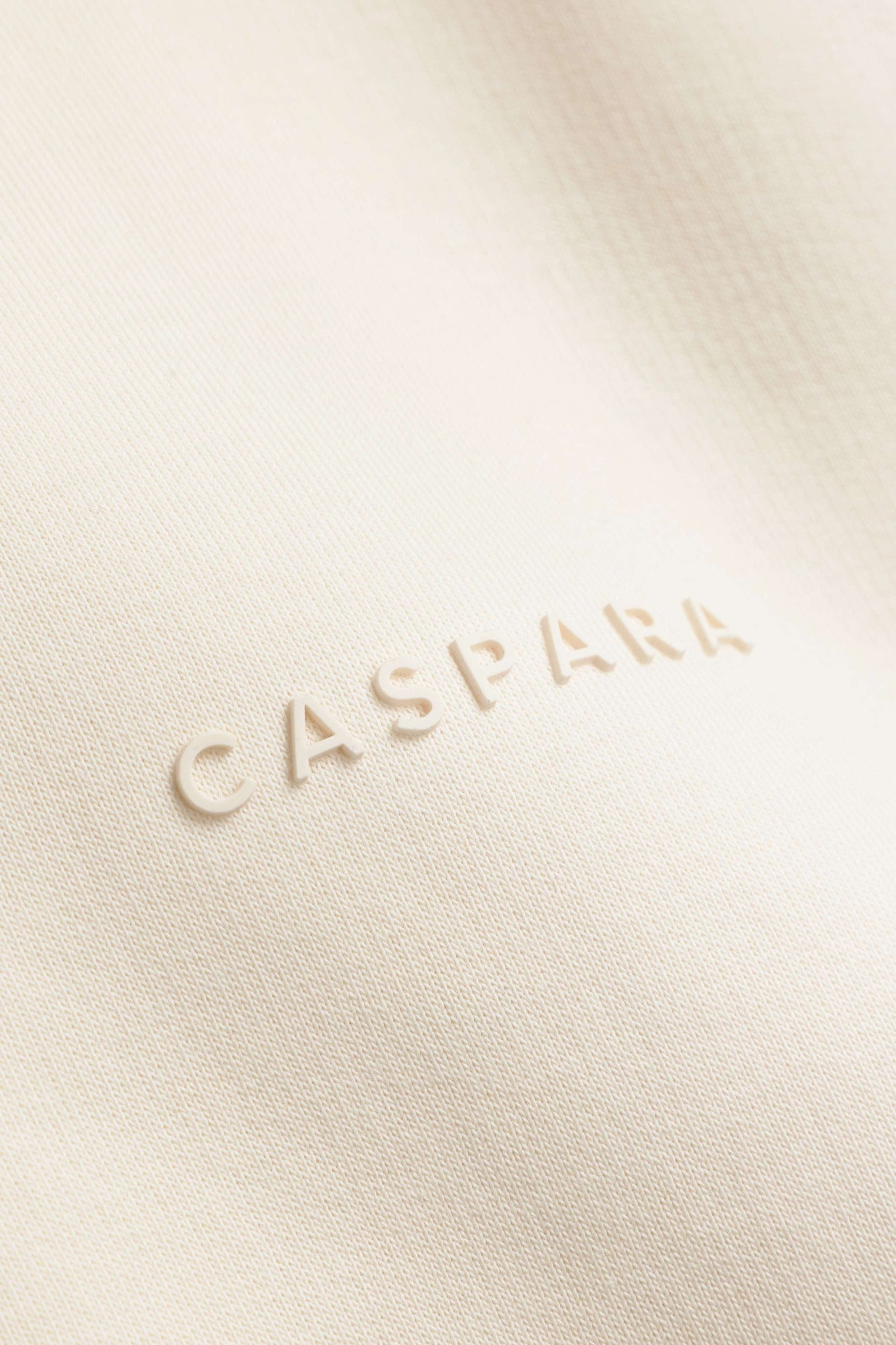 Close-up of the tone-on-tone rubber CASPARA logo on the creme Cosmo zip hoodie.